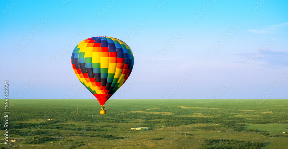 Obraz premium A colorful air balloon is flying in free flight over the field. Bird's-eye view. Multi colored balloon in the blue sky
