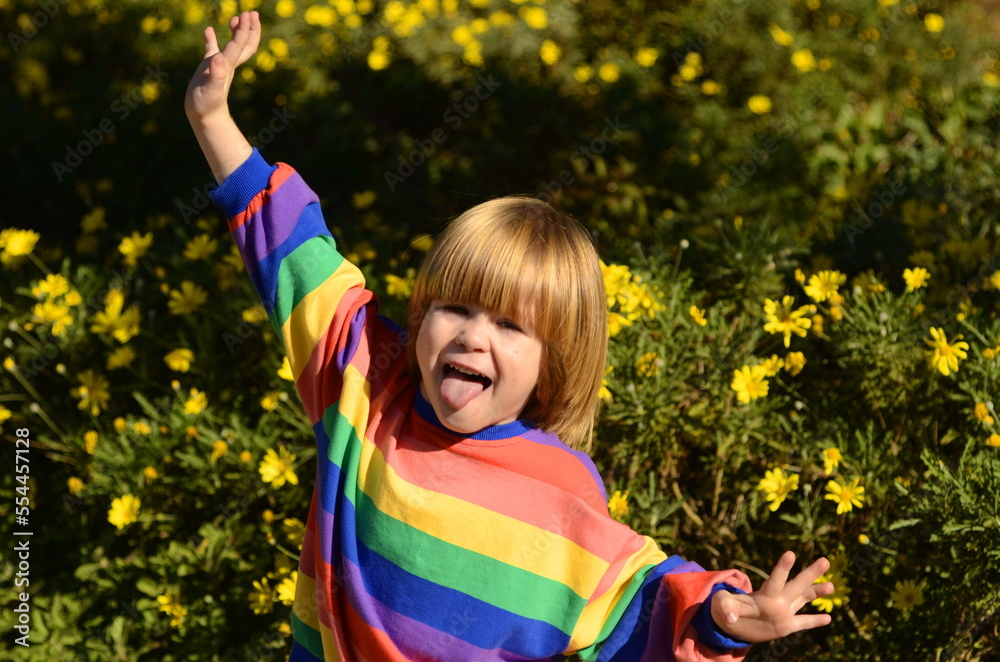 A positive boy, a funny child rejoices against the background of yellow flowers. Bright mood and rainbow T-shirt. Color emotions, joy. Space for text. The little boy smiles, hands up and laughs.