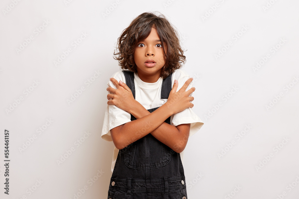 Stupefy brunette young guy in denim overalls keeps arms crossed, feels uncomfortable and scared, poses over white background Stupefy brunette young guy in denim overalls keeps arms crossed, feels uncomfortable and scared, poses over white background