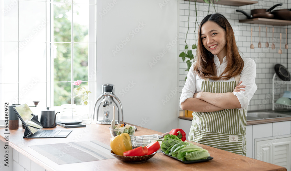 Portrait of young happy woman wearing appron standing with arms crossed in the kitchen room, prepares cooking healthy food from fresh vegetables and fruits. Portrait of young happy woman wearing appron standing with arms crossed in the kitchen room, prepares cooking healthy food from fresh vegetables and fruits.