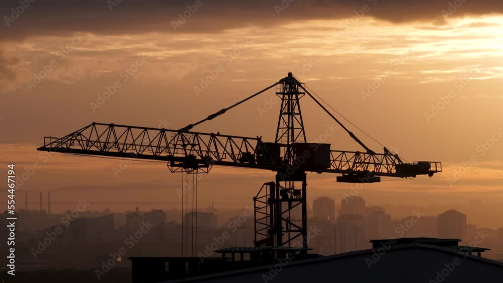 Silhouette of tall construction crane that turns arrow to lift delivery of building materials. Work on construction site. Against background of sunset and orange sunset in the city. Silhouette of tall construction crane that turns arrow to lift delivery of building materials. Work on construction site. Against background of sunset and orange sunset in the city.