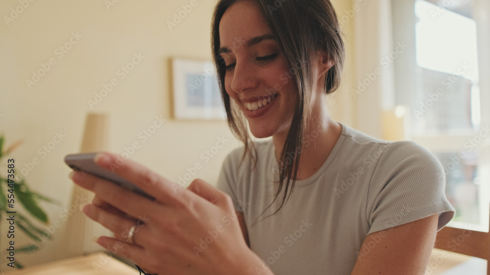 Smiling young woman scrolling through photo on her mobile phone while sitting in the kitchen Smiling young woman scrolling through photo on her mobile phone while sitting in the kitchen
