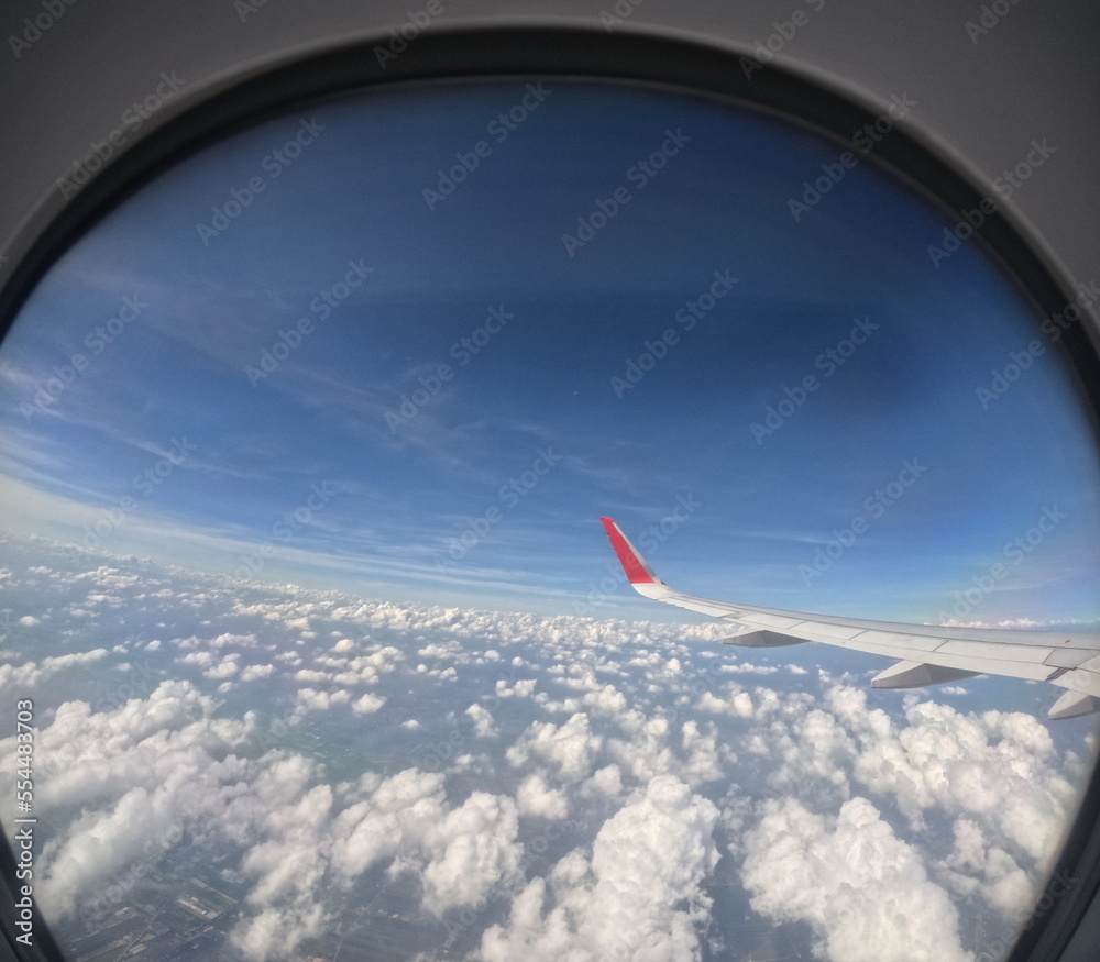 looking through the window of a passenger plane Plane wing tips, bright blue skies, dense white clouds, and the ground can be seen in the distance.
