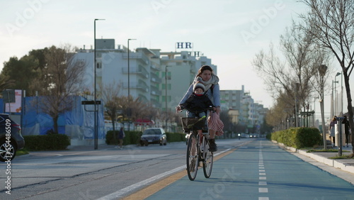 Mother rides bicycle with child in front in bike lane by sea shore. Parent riding bike outdoors during cold day