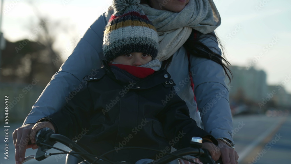 Mother rides bike with child in front during cold winter day with sunlight shining in background. Mom riding bicycle
