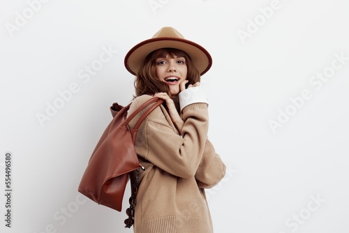 an excited woman posing on a light background in a shirt, sweater and hat leaned forward a little holding a bag behind her back smiling cheerfully
