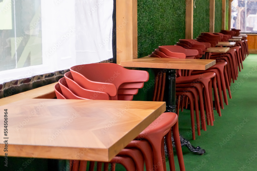 Red plastic patio chairs and square wooden tables are stacked against a ...