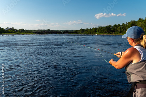 A middle age female stands in a large salmon river casting a fishing rod holding line in her left hand. The blonde long haired lady is wearing a blue hat, white t-shirt, and waders fly-fishing. 
