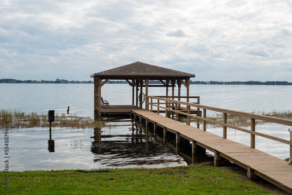 A wooden pier with a large square hut at the wharf. The platform marina has a wooden rail around the wharf and a metal roof. The gazebo stage is over a blue calm ocean. The sky is blue with some cloud A wooden pier with a large square hut at the wharf. The platform marina has a wooden rail around the wharf and a metal roof. The gazebo stage is over a blue calm ocean. The sky is blue with some cloud