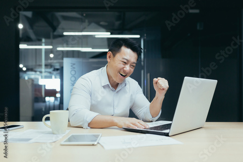 A young Asian man sits in the office at the table, rejoices at the victory and the win, looks at the laptop, participates in the auction, places bets. He smiles, shows a yes gesture with his hand