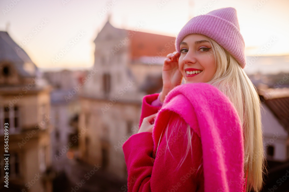 Happy smiling fashionable blonde woman wearing trendy knitted beanie hat, woolen scarf, coat, posing outdoor, with beautiful city view. Selective focus. Copy, empty space for text