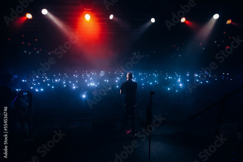 Popular singer in front of crowd on scene in night club. Bright stage lighting, crowded dance floor. Phone lights at concert. Band blue silhouette crowd. People with cell.