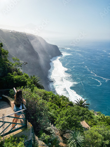 A young tourist woman stands at the viewpoint of a cliff overlooking the ocean in Tenerife, Canary Islands