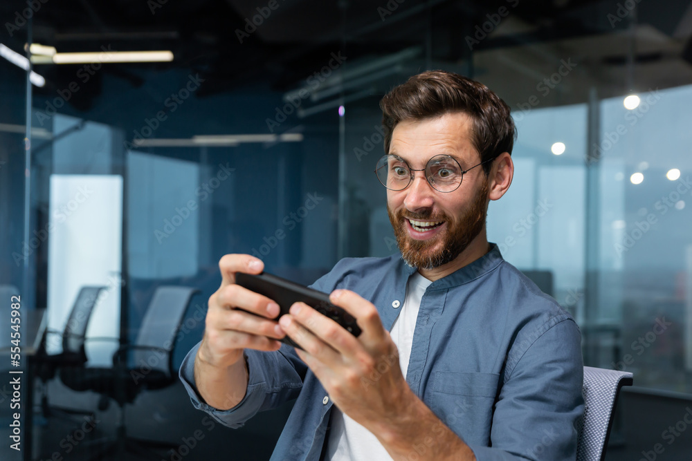 Mature businessman with beard is playing game on smartphone, man is having fun during break at workplace, businessman inside modern office. Mature businessman with beard is playing game on smartphone, man is having fun during break at workplace, businessman inside modern office.