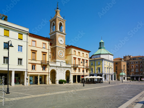 Piazza Tre Martiri Three Martyrs square with traditional buildings with clock and bell tower in old historical touristic city centre Rimini with blue sky background, Emilia-Romagna, Italy