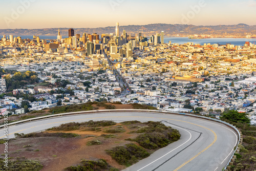 San Francisco skyline warmly lit by a setting sun in autumn. A winding road climbing Twin Peaks is in foreground.