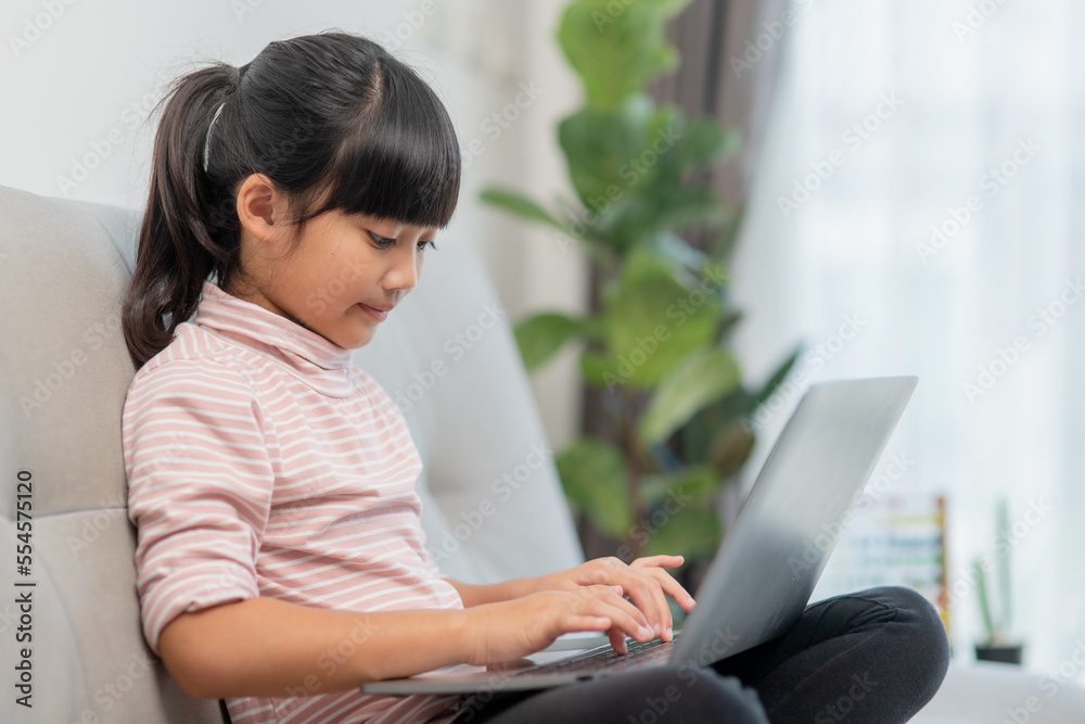 Asian Little school kid girl use laptop computer sitting on sofa alone at home. Child learning reading online social media content, play education lessons game chatting with friends. Asian Little school kid girl use laptop computer sitting on sofa alone at home. Child learning reading online social media content, play education lessons game chatting with friends.