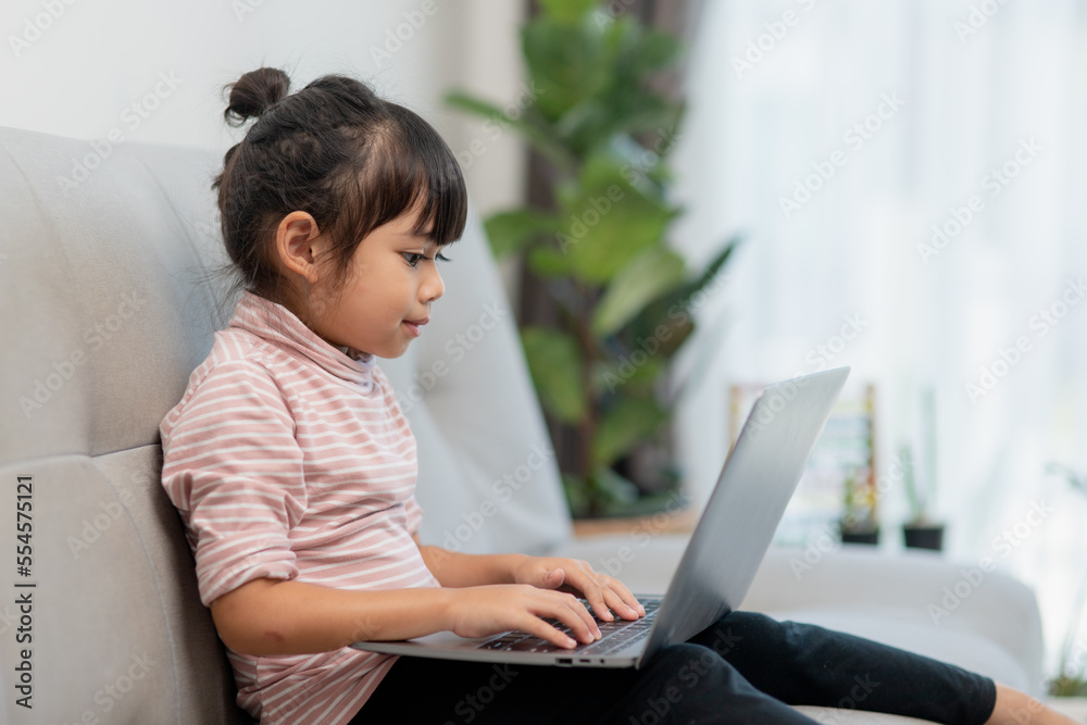 Asian Little school kid girl use laptop computer sitting on sofa alone at home. Child learning reading online social media content, play education lessons game chatting with friends. Asian Little school kid girl use laptop computer sitting on sofa alone at home. Child learning reading online social media content, play education lessons game chatting with friends.