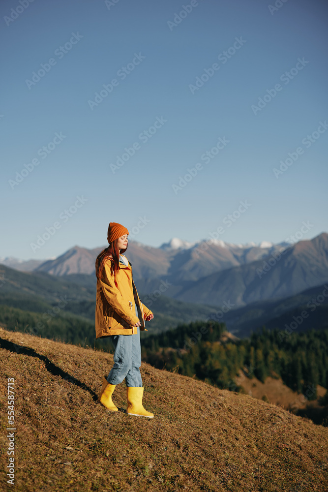 Fototapeta premium Woman autumn full-length walk on the hill smile with teeth and look at the mountains in a yellow raincoat and jeans happy hiking trip, freedom lifestyle 