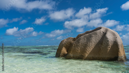 A huge granite boulder with folded slopes rises in the ocean. Clear turquoise water and blue sky with clouds. Seychelles. La Digue island. Anse Source D’Argent beach 