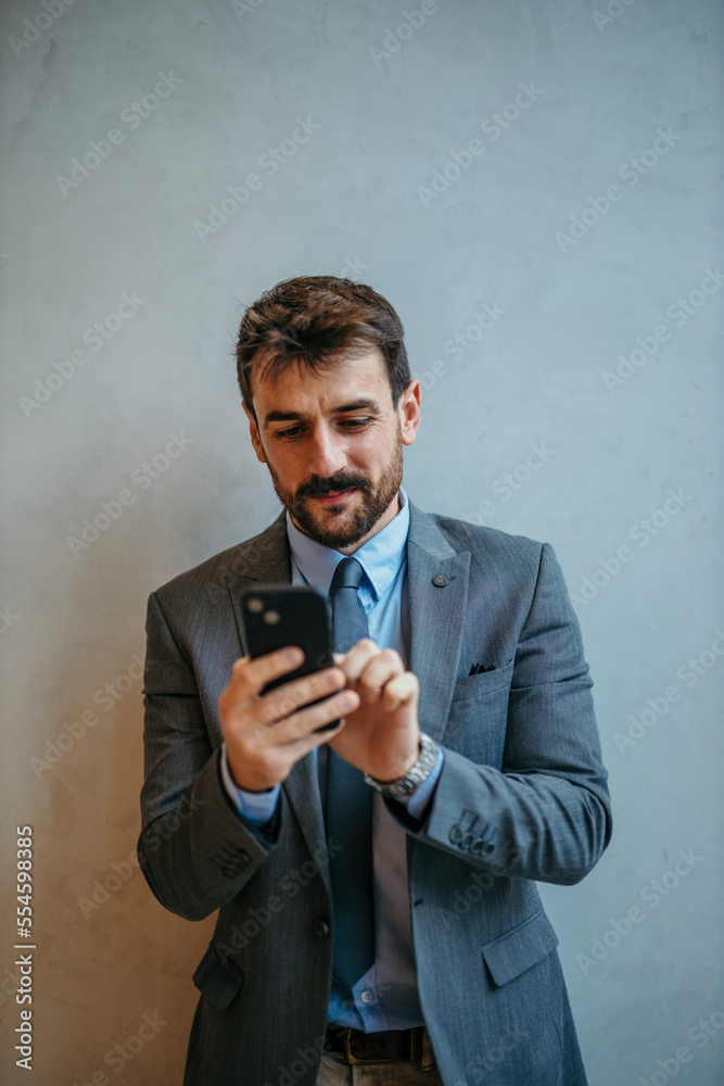 Handsome smiling businessman standing in a gray suit, holding and using a mobile phone isolated on a white wall. Handsome smiling businessman standing in a gray suit, holding and using a mobile phone isolated on a white wall.
