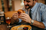 A handsome businessman in a blue shirt and tie with rolled up sleeves is eating a burger and french fries, and drinking a beer after work in a restaurant.