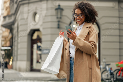 Fashion afro american woman after shopping with paper bags standing in city and using phone photo