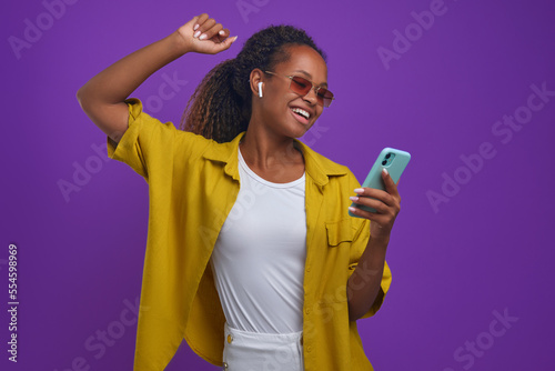 Tableau sur toile Young dancing cheerful African American woman with wireless earphone holds mobil