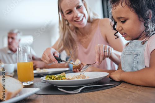 Family  mother and cutting food for girl while having lunch at dinner table in home. Love  foster care and happy mom helping child with eating  smiling and enjoying a delicious meal together in house