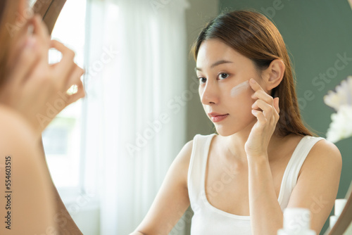 Fresh skin, beautiful smile of asian young woman, girl looking at mirror, hand applying moisturizer lotion on her face, putting cream treatment before makeup cosmetic routine at home. Facial Beauty. © KMPZZZ