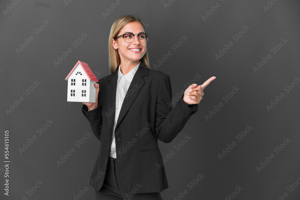 Blonde Uruguayan girl holding a house toy isolated on black background pointing finger to the side and presenting a product Blonde Uruguayan girl holding a house toy isolated on black background pointing finger to the side and presenting a product