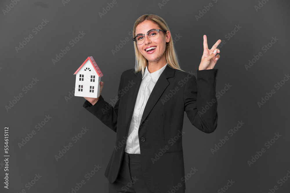 Blonde Uruguayan girl holding a house toy isolated on black background smiling and showing victory sign Blonde Uruguayan girl holding a house toy isolated on black background smiling and showing victory sign