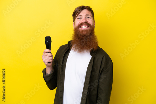 Redhead man with beard picking up a microphone isolated on yellow background smiling a lot