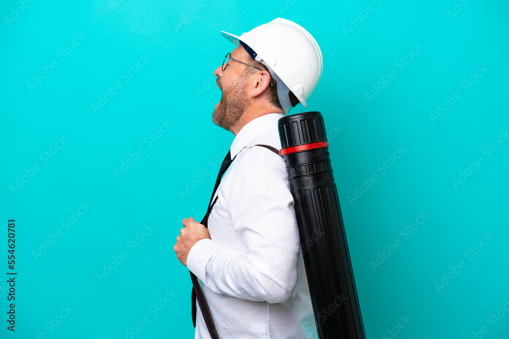 Middle age architect man with helmet and holding blueprints isolated on blue background laughing in lateral position Middle age architect man with helmet and holding blueprints isolated on blue background laughing in lateral position