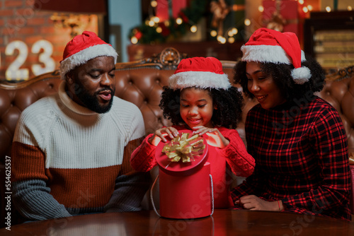 Happy African American family celebrating Christmas mother, father and adorable daughter opening Christmas gift box in leaving room with Christmas decorations at home.