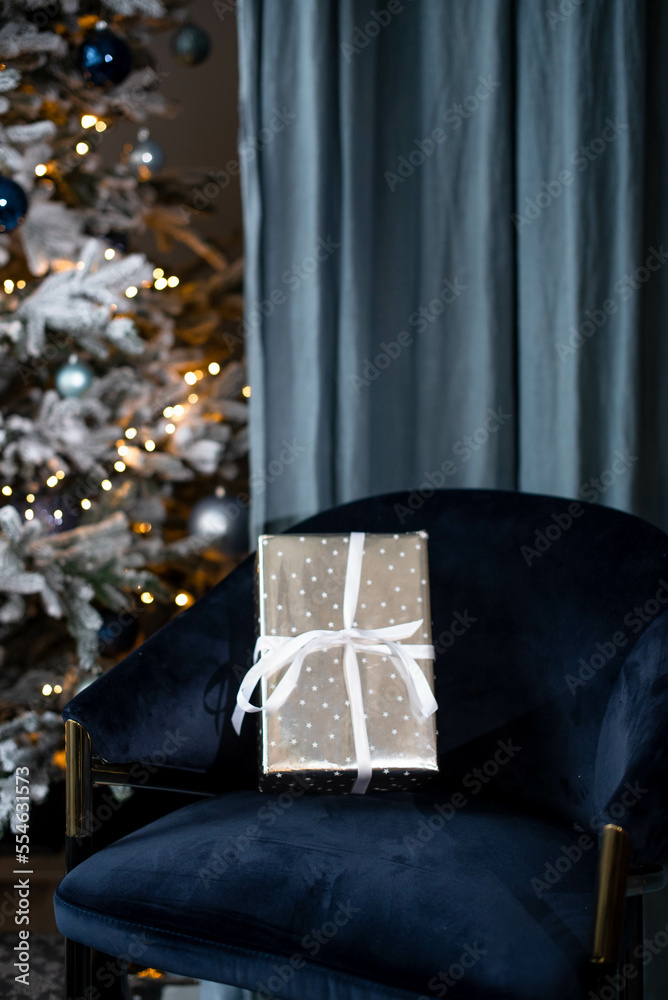 A gift in a silver wrapper with a white ribbon on a velvet blue chair against the background of a Christmas tree..