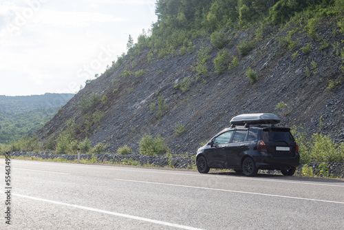 Black hatchback car with an autobox on the roof, with a trunk on the roof of the car, stands along the highway in the middle of the mountains.The concept of auto travel,adventure,adventurism,lifestyle