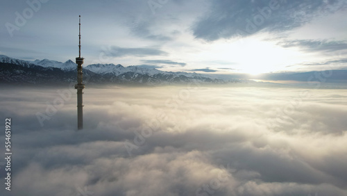 The TV tower looks out of the clouds at sunset. Top view from a drone on a double layer of clouds. Yellow-orange color from the rays of the sun. Shadows on clouds. High mountains in the distance