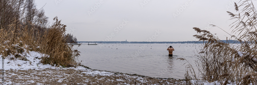 Ein junger Mann geht mit im Winter Badehose und Mütze in einem See schwimmen, Panorama