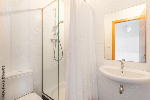 Small simple bright bathroom with shower area enclosed by glass partition and screen. Square mirror above washbasin shows front door to bedroom. There is white toilet against wall.