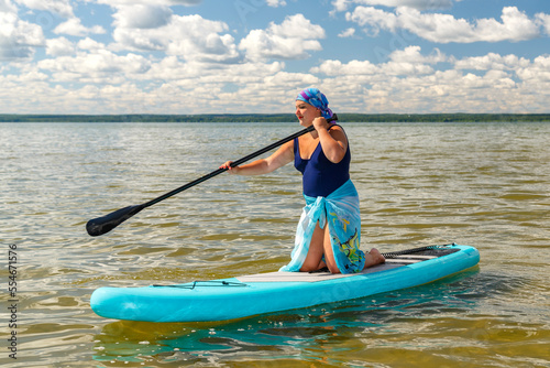 A Jewish woman in a kerchief kisuy rosh in a pareo on her knees on a SUP board swims in the lake on a sunny day.