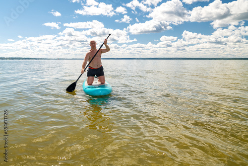 A man balances on a SUP board with a paddle in the lake on a sunny day against the backdrop of white clouds.