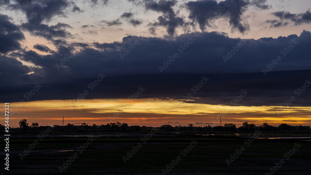 Scenery of thick clouds floating above the silhouette of trees and rice fields.