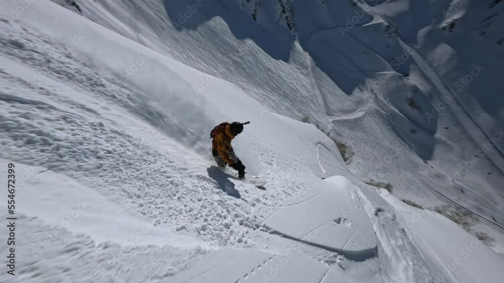 Male snowboarder freeride riding from mountain summit drop snow splash extreme sport tourism aerial view. FPV drone shot active man snowboard rider enjoy adrenaline freedom winter cliff landscape 4k