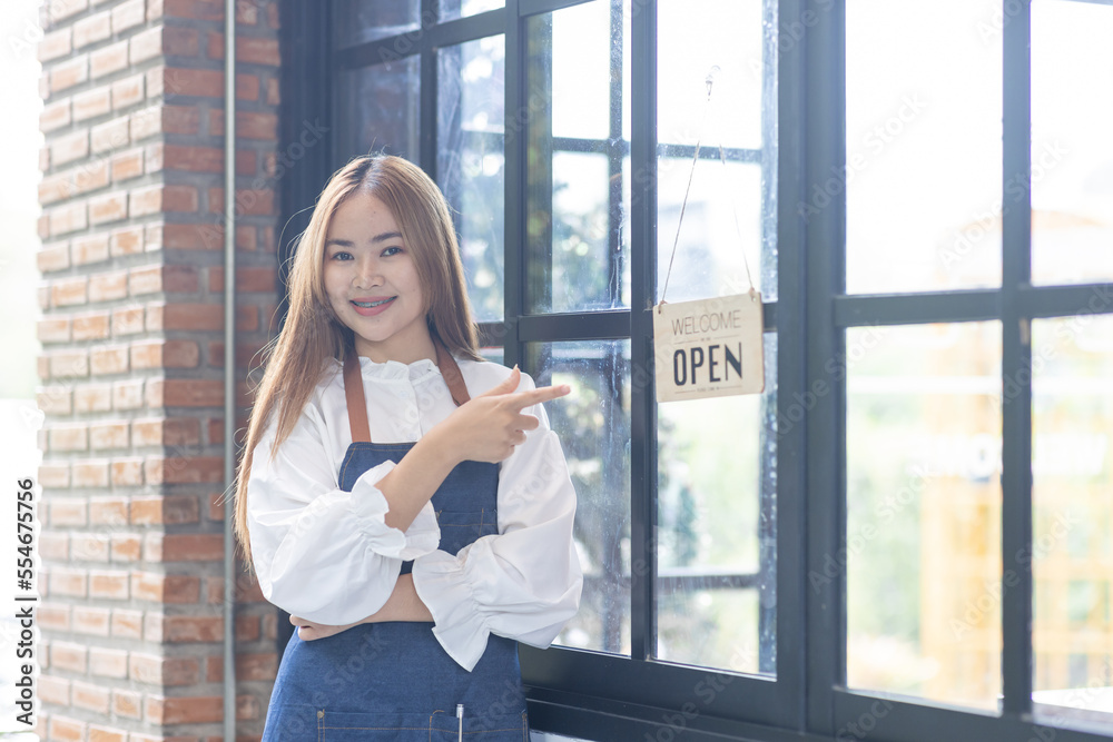 Portrait of happy waitress standing at restaurant entrance. Portrait of young business Asian woman attend new customers in her coffee shop. Smiling small business owner showing open sign in her shop.