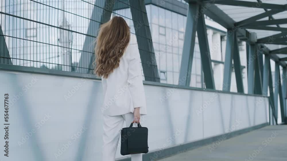 Young confident business woman in white suit walking with bag of laptop. Busy beautiful girl leaving office building after work. Slow motion Young confident business woman in white suit walking with bag of laptop. Busy beautiful girl leaving office building after work. Slow motion
