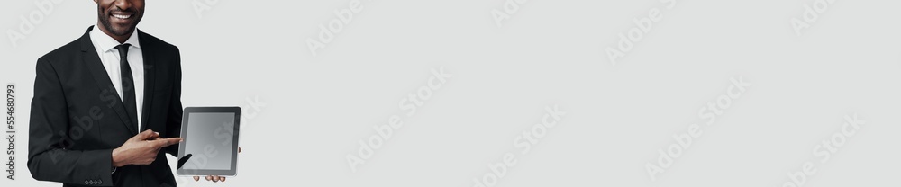 Confident young African man in formalwear pointing copy space on digital tablet while standing against grey background Confident young African man in formalwear pointing copy space on digital tablet while standing against grey background