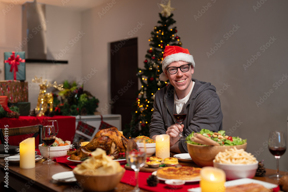 Happy man in glasses wear red Santa Claus hat holding glass of wine while celebrating Christmas dinner with their family.