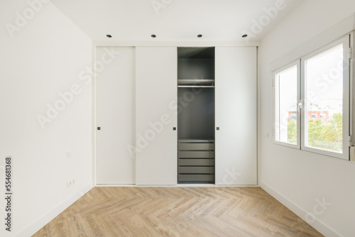 Room with a custom built-in wardrobe with white sliding doors  white painted smooth walls  a gray wardrobe interior with drawers and an oak floor
