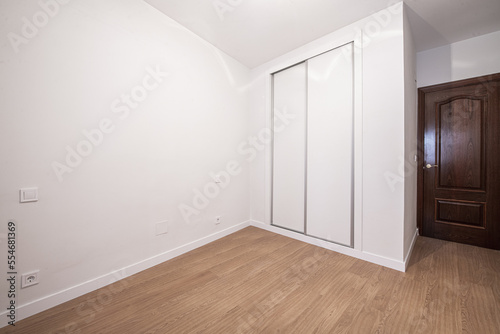 A room with a custom built-in wardrobe with white sliding doors, white painted smooth walls and dark wooden doors